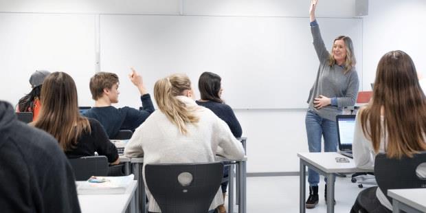Bilde av  elever som sitter med ryggen til ved pultene sine i et klasserom. En lærer står foran elevene og snakker til klassen.