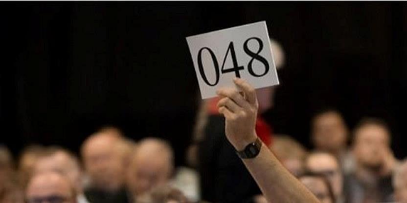 Bilde av en gruppe mennesker som sitter ved ulike bord. En løfter et skilt som det står 048 på. Illustrasjon på årsmøte