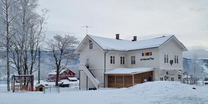 Hvitmalt barnehage, tatt utendørs, med mye snø, fjell i bakgrunnen, og noen trær og en lekeplass. 