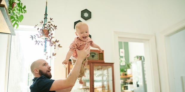 Baby og far gjør løftende barnefly i luften på sofaen i en familiestue. Mannlig person eller pappa med et småbarn for kvalitetstid, kjærlighet og lykke mens de leker sammen.