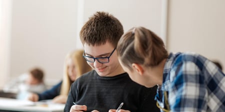 Kautokeino barne- og ungdomsskole, Nærbilde av elev og kvinnelig lærer ved pulten i klasserommet. Bøker på pulten, De smiler, Eleven får hjelp av læreren, Støttelærer