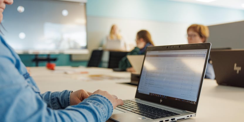 Bilde av en bok støttet opp mot en bærbar pc, bakgrunnen er i klasserom med tavler.