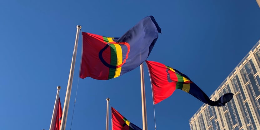 Samiske flagg blafrer i vinden. Det er solskinn og et
moderne bygg sees i bakgrunnen.