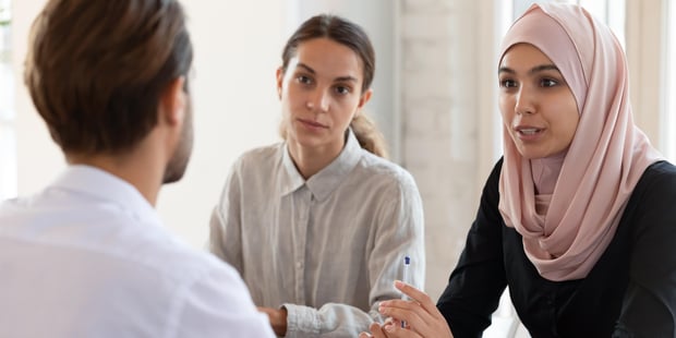 To yngre kvinner og en yngre mann sitter ved et møtebord. Den ene kvinnen har hijab. Mannen sitter med ryggen til.