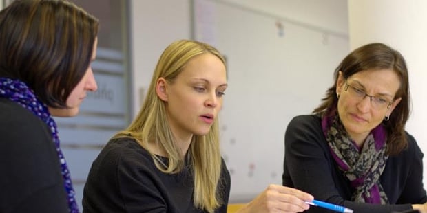 3 personer som ser på en bok som personen i midten peker på med en penn. Foto.
