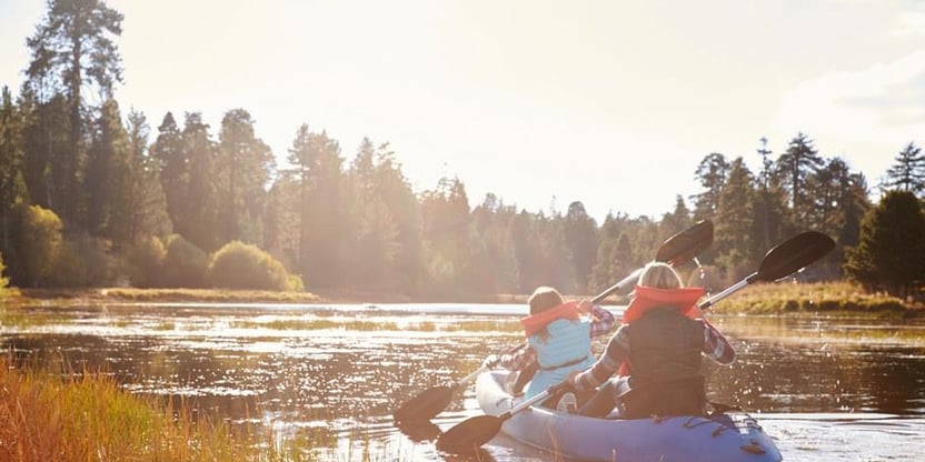 To personer padler i en kano på et vann. Sol, skog i bakgrunnen. Foto.