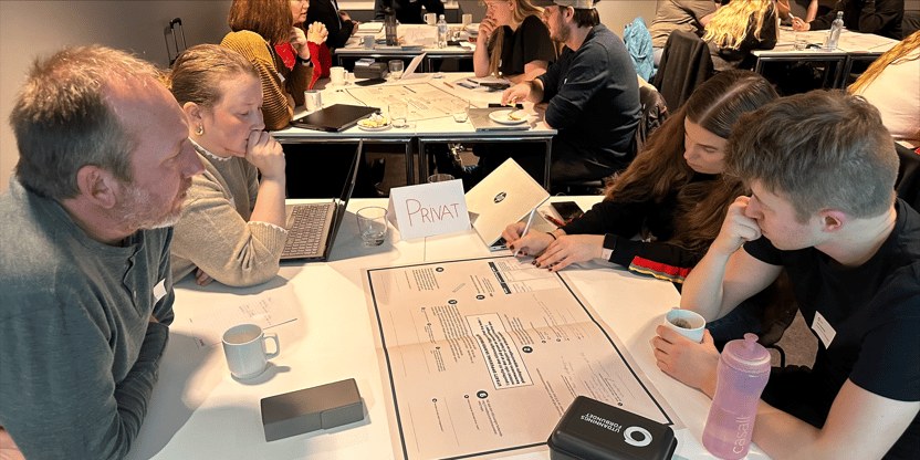 Fire arbeidsplasstillitsvalgte sitter  rundt et bord og jobber med caser på en dialogduk. Et brettekort fastslår at de jobber i det private. Eller på bordet står et glass, en kaffekopp, en drikkeflaske og en matboks med Utdanningsforbundets logo. En av de tillitsvalget, kvinne, skriver på duken, og en annen holder hånde mot kinnet, Kvinne, en har armene foldet mot brystet og lener seg på bordet, mann, og den siste holderen hånd mot kinnet mens den ander holder en kaffekopp, mann. I bakgrunnen sees andre tillitsvalgte som også jobber.