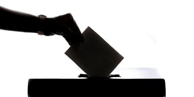 Svart-hvit silhuett av hånd som putter stemmeseddel ned i en boks. Foto.
