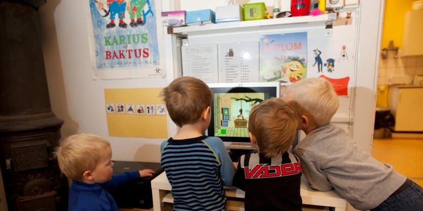 Fire barnehagebarn står med ryggen til fotografen foran en dataskjerm. De spiller trolig et dataspill.
