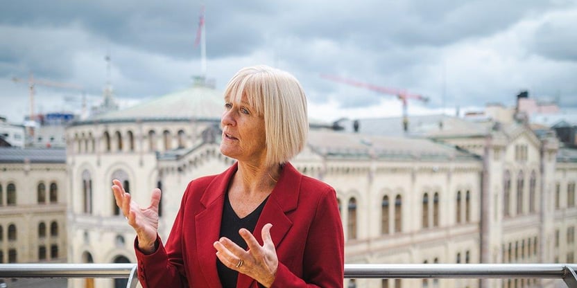 Kvinne i rød blazer står ute på balkong og gestikulerer med armene