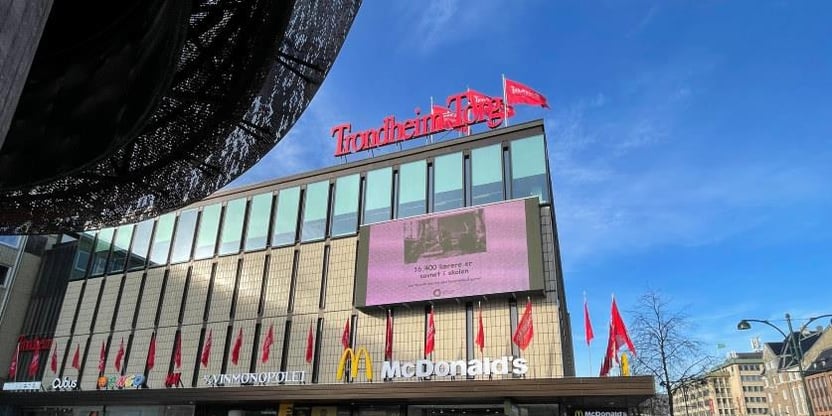 Bilde fra Trondheim Torg, fasade kjøpesenter. Butikklogoer. Folk går forbi i forgrunnen. Billboard på fasaden viser kampanjen «Savnet Lærer» «16 400 lærere er savnet i skolen». Foto.
