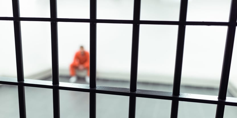 Fra et fengsel med en innsatt som sitter på en benk i oransje fangedrakt bak et gitter.
