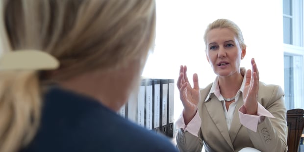 En dame med oppsatt hår, snakker mens hun ser på en annen person som sitter med ryggen til. Hun har hendene løftet som for å forklare noe.