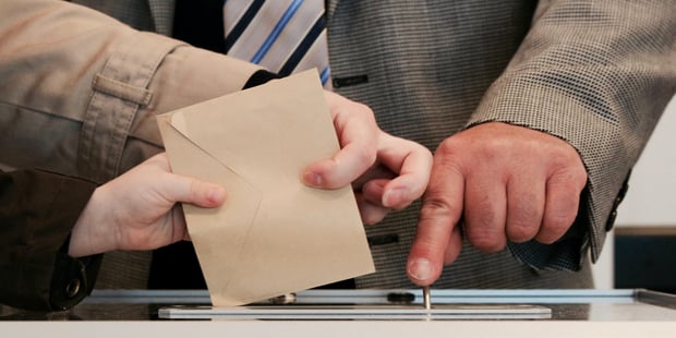 Bildet viser hender som holder en konvolutt, som skal puttes i en urne, og en hånd med finger på en knapp som åpner urnen. Bildet illustrerer en stemmegivning i valg.