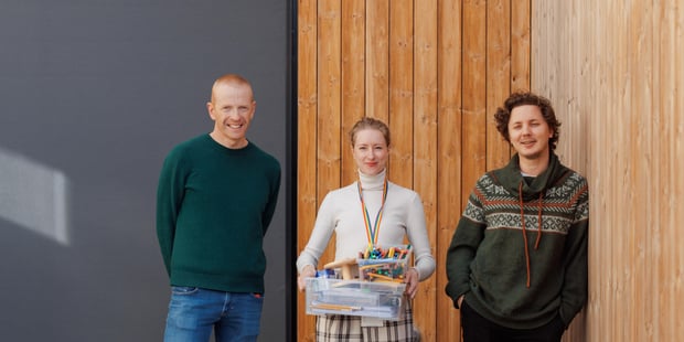 2 menn og en kvinne står oppreiste, og ser mot fotografen. Kvinnen holder en kasse med lekesaker og tegnesaker i hendene.