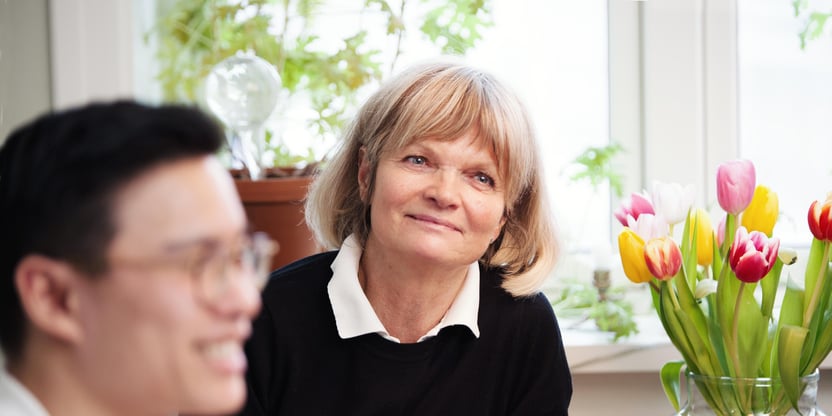 Ung mann og eldre kvinne sitter foran pc ved kjøkkenbord. Det er tulipaner på bordet.