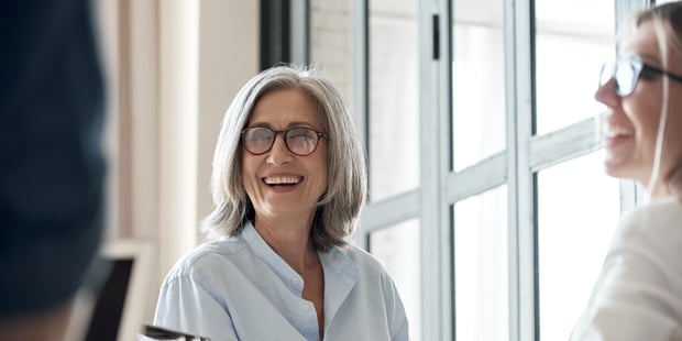 En dame med halvlangt, grått hår og briller smiler med åpen munn mens hun og en annen person ser på en tredje person.