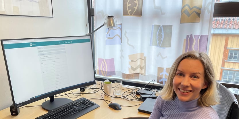 Bilde av Kristine smilende foran skjermen på kontoret.