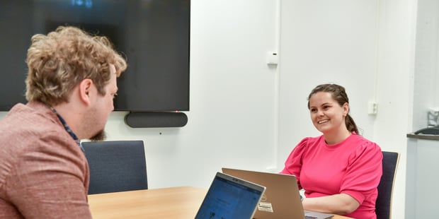 En kvinne og en mann sitter på hver sin side av ved side et bord med hver sin PC. Vi ser ansiktet til kvinnen, profilen til mannen. Foto.