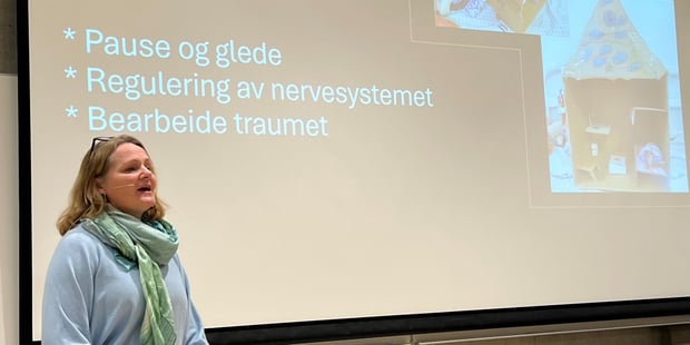 Kvinne står foran storskjerm og forteller. Skjermbildet sier "Lek som psykologisk kinderegg".
