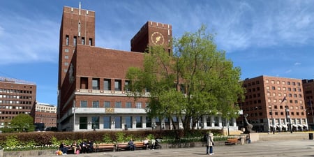Oslo rådhus fotografert fra sjøsiden. Det er vår og blå himmel. Et tre står med lysegrønt bladverk foran rådhuset.
