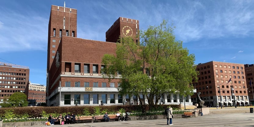 Oslo rådhus fotografert fra sjøsiden. Det er vår og blå himmel. Et tre står med lysegrønt bladverk foran rådhuset.