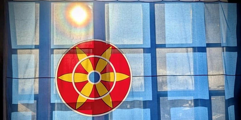 Bilde av stort kvenflagg foran vinduer med sol som skinner igjennom i overkant av solrosen i flagget.