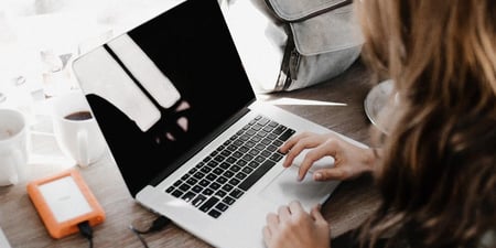Bildet viser en dame som sitter og skriver på en laptop. Du ser damen fra, og hele tastaturet.