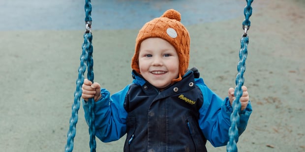 Gutt sitter på en bildekk-huske, ute i en barnehage, har på seg lue og yttertøy