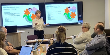Kvinnelig foredragsholder som står foran ei forsamling. PP-presentasjon på lerret i bakgrunnen