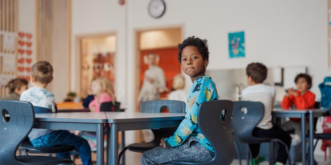 Bildet viser en elev ved Oksenøya barneskole. Han sitter ved pulten og har snudd seg mot fotografen. Han ser lett undrende ut. I bakgrunnen skimtes mange klassekamerater.