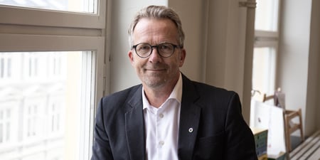 Portrettbilde av mann i hvit skjorte og dressjakke som smiler til kamera. Han sitter i vinduskarmen.