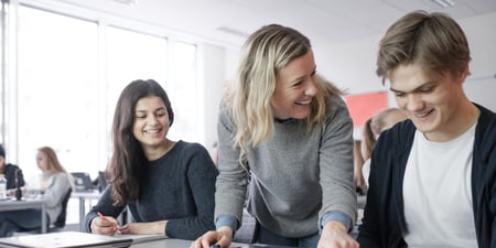 Bildet viser en blid lærer som står ved en pult, hvor det sitter to smilende elever.