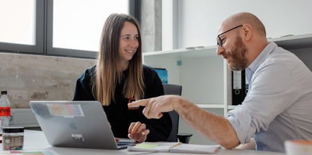 To lærere på arbeidsrom som sitter ved et bord. Den ene mannlige læreren peker på PC ’n til den kvinnelige læreren. Det er god stemning mellom dem.