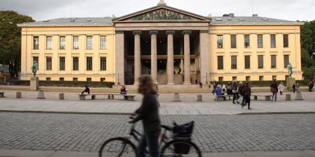 Universitetet i Oslo,  juridisk fakultet. Bygningen er i klassisk stil med store søyler. Det er sol og flere fotgjengere og en sykelist beveger seg på fortau og gate foran bygget.