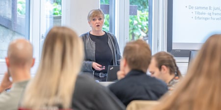 Lærer i et klasserom står og snakker til en gruppe voksne som sitter med ryggen til fotografen.