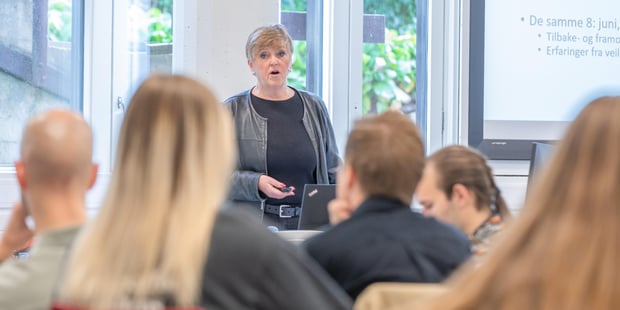Lærer i et klasserom står og snakker til en gruppe voksne som sitter med ryggen til fotografen.