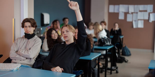 Flere elever sitter ved pultene i et klasserom. En av dem rekker opp hånden. Illustrasjonsfoto