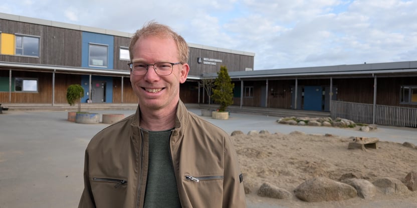 Halvnært bilde av en mann som står foran et barnehagebygg og smiler.