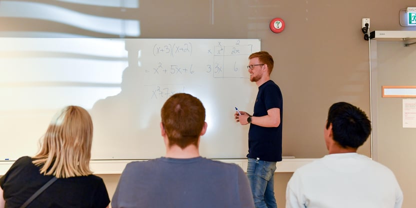 En mann står foran et whiteboard med en tusj i hånden. Tre personer sitter på hver sin stol og ser på mens han forklarer noe.