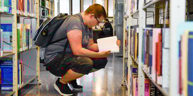 En student sitter på huk i et universitetsbibliotek og leser i en bok. Illustrasjonsfoto