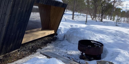 Bilde av en gapahuk i tre, med en bålpanne og en trebenk som begynner å tine fra, fra snøen. I bakgrunnen er det furutrær.