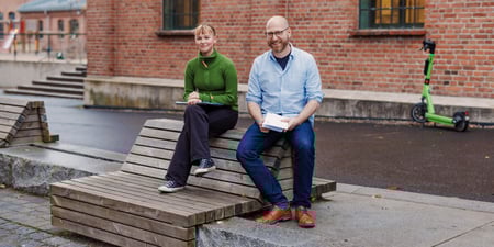 En dame og en mann sitter på en benk i en skolegård. De smiler. Foto.