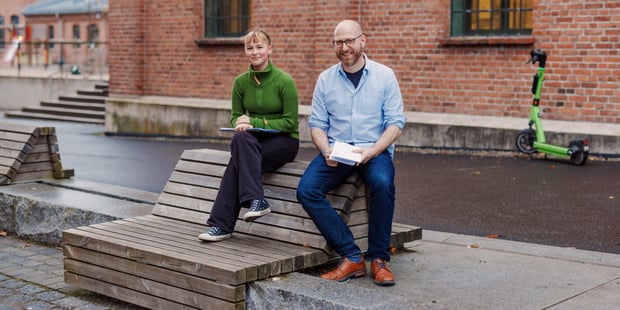En dame og en mann sitter på en benk i en skolegård. De smiler. Foto.