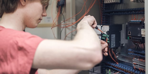 En elev på yrkesfag skrur med en skrutrekker inni et elektrisk skap med sikringer og ledninger. Illustrasjonsfoto