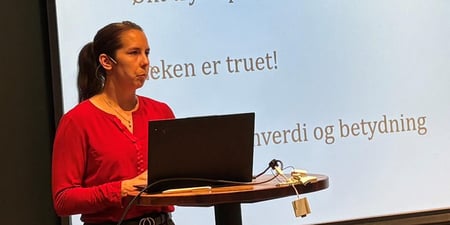 Sentralstyremedlem Linn Therese Myhrvold med pc på forelesningsbod foran seg og skjerm med nytt fra sentralstyret i bakgrunnen holder foredrag på lokallagsleder og hovedtillitsvalgtsamling.