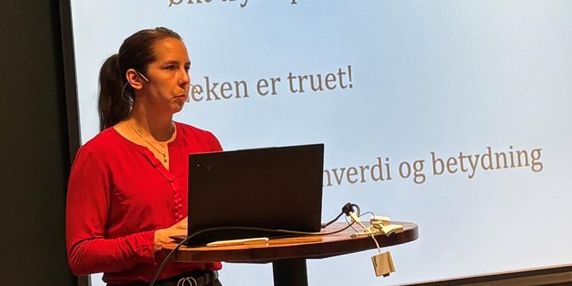 Sentralstyremedlem Linn Therese Myhrvold med pc på forelesningsbod foran seg og skjerm med nytt fra sentralstyret i bakgrunnen holder foredrag på lokallagsleder og hovedtillitsvalgtsamling.