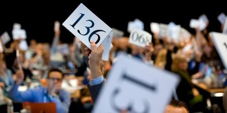 Mange personer i en stor møtesal som voterer i en sak. Foto.