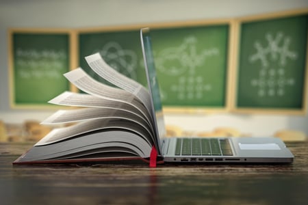 Bilde av en bok støttet opp mot en bærbar pc, bakgrunnen er i klasserom med tavler.