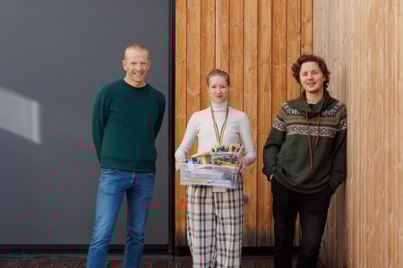 2 menn og en kvinne står oppreiste, og ser mot fotografen. Kvinnen holder en kasse med lekesaker og tegnesaker i hendene.